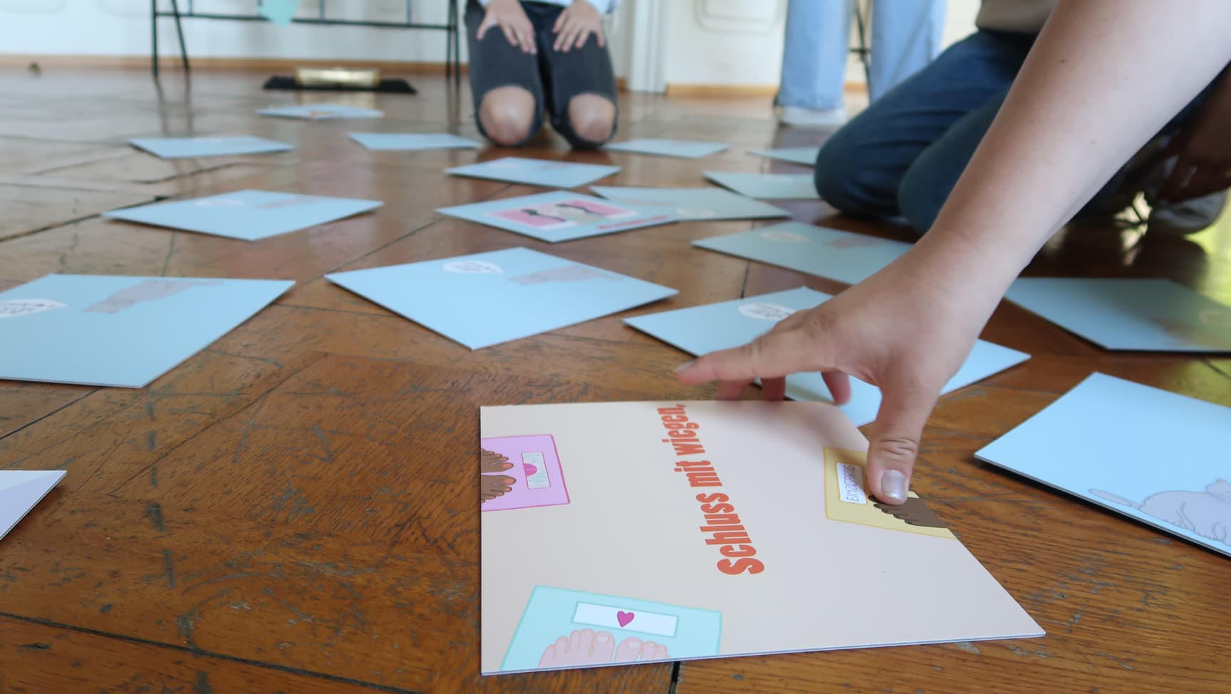 Auf dem Boden liegen große Memorykarten. Personen sitzen im Kreis um die Karten, eine Person greift nach einer Karte auf der steht: schluss mit wiegen