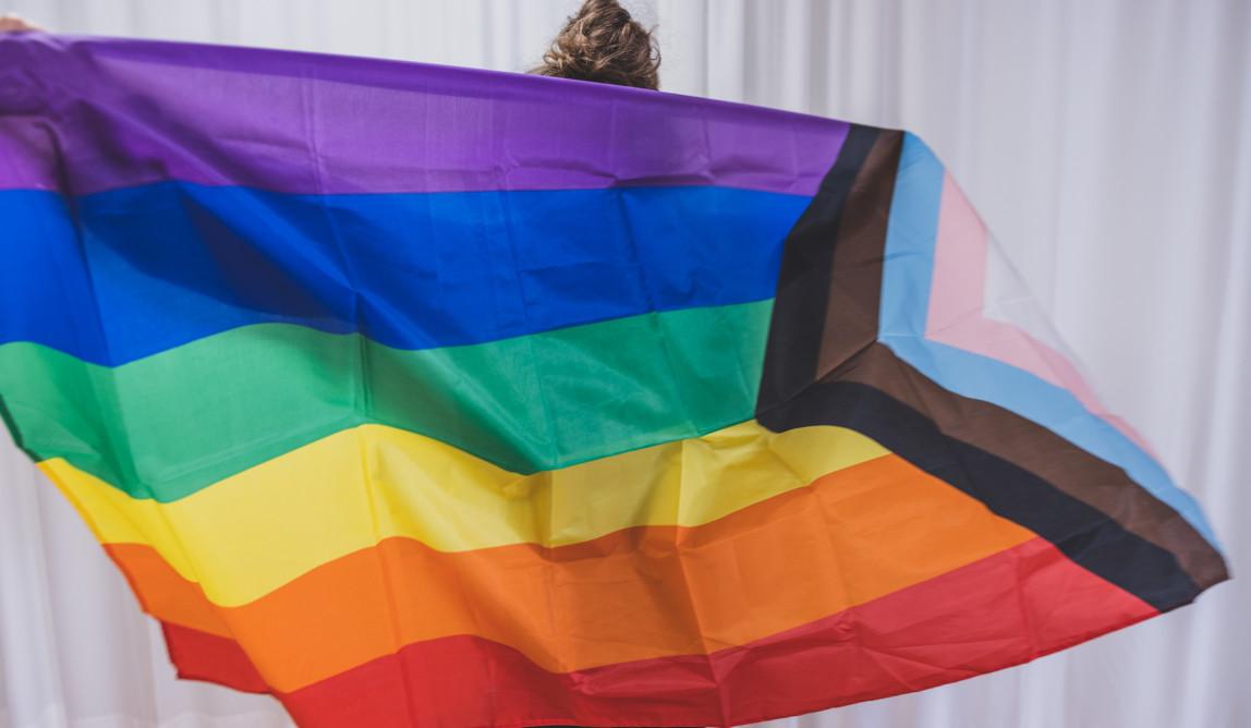 Eine Person hält eine große Progress Pride Flag mit beiden Armen weit ausgestreckt vor sich, das Gesicht ist nicht zu sehen.