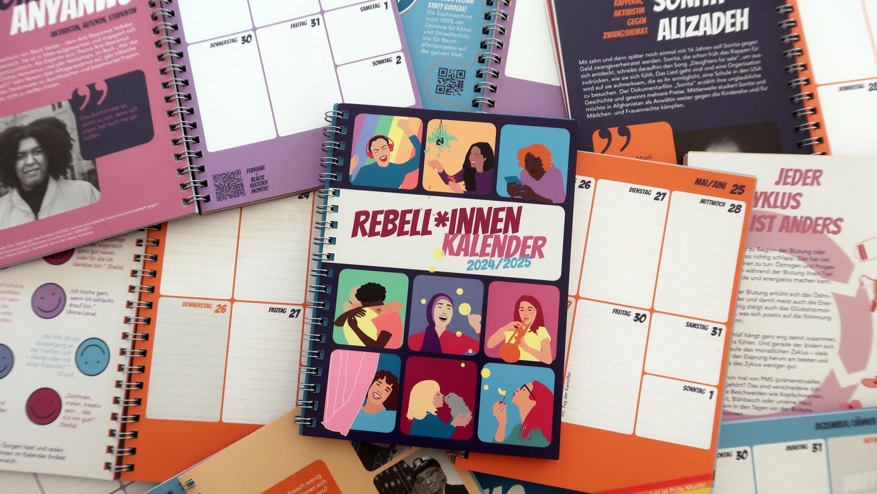 Der Rebell*innen Kalender 2024/25 liegt inmitten vieler aufgeschlagener anderer Kalender.