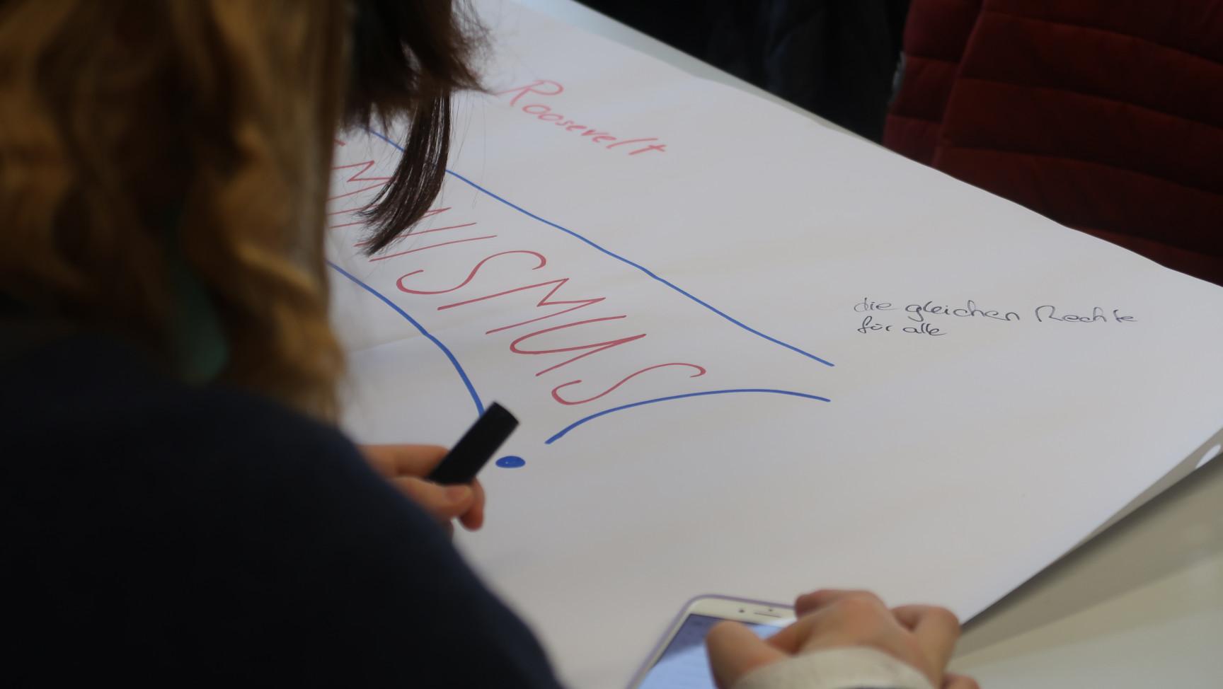 Detailaufnahme über die Schulter einer Person, die ein Stift und Smartphone hält. Im Hintergrund ist ein Teil eines Plakats sichtbar.