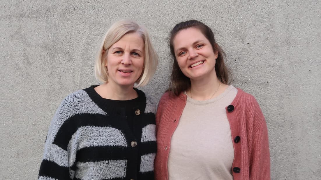 Zwei Frauen stehen vor einer grauen Wand und lachen in die Kamera.