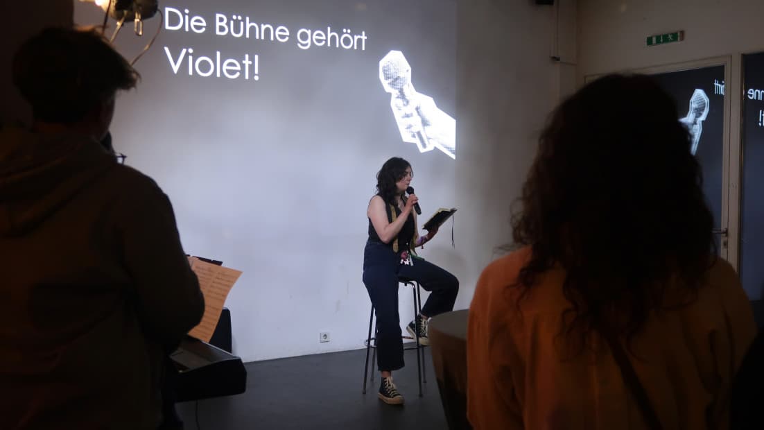 Eine junge Person sitzt auf einem Barhocker und liest aus einem Notizbuch. Im Hintergrund steht an die Wand projiziert "Die Bühne gehört Violet"