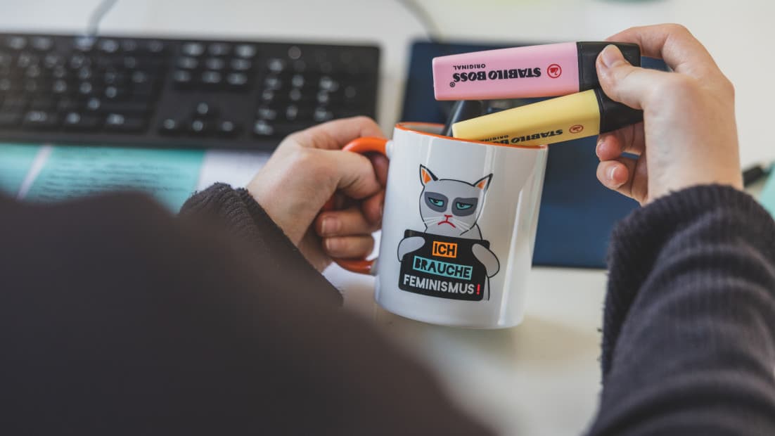 Jemand nimmt zwei Stifte aus einem Becher mit der Aufschrift "Ich brauche Feminismus!"