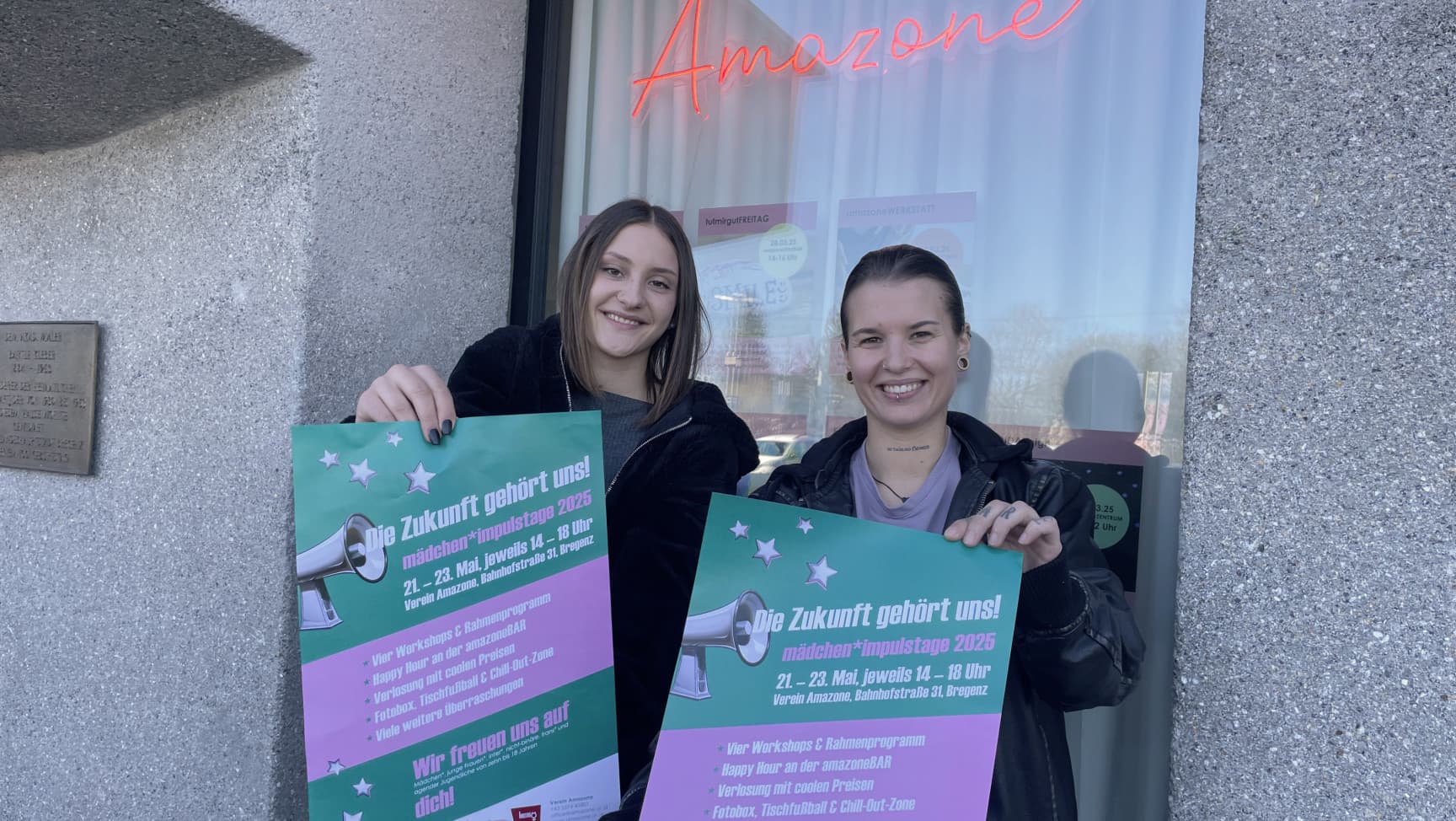 Zwei junge Frauen* stehen vor einem Fenstern mit dem Schriftzug Amazone und halten Plakate.